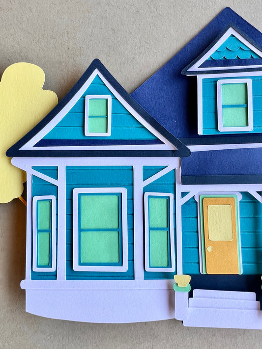 Closeup of a paper blue house with green windows and a yellow door on a beige background